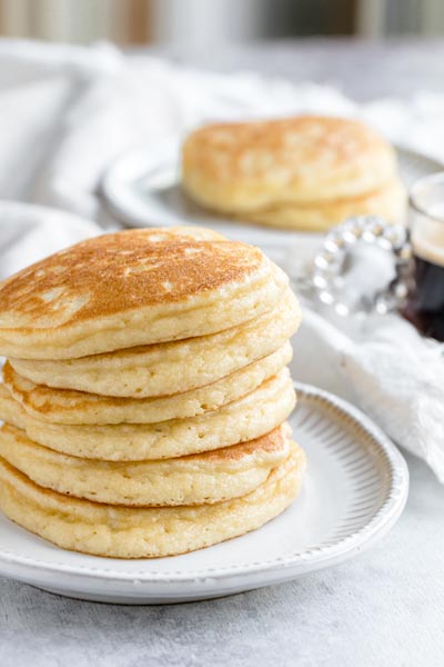 A stack of plain pancakes on a plate with a cup of coffee to the side.