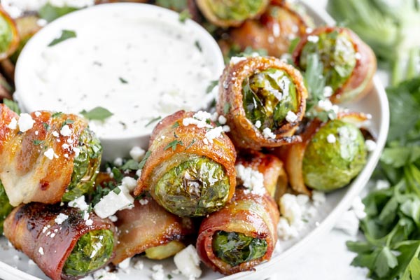 bacon wrapped brussels sprouts it in a serving tray with a bowl of ranch dressing in the center and topped with crumbled feta cheese