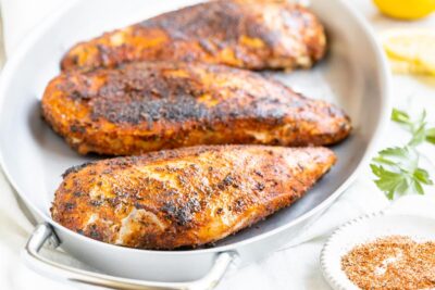 Juicy blackened chicken breasts on a stainless steel skillet with handles next to a Cajun blend of spices in a dish and parsley.