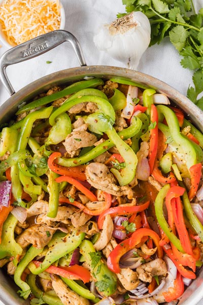 A skillet filled with chicken fajitas next to shredded cheese and garlic.