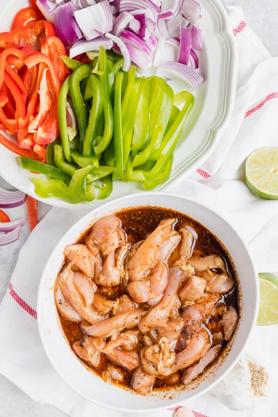 A bowl with raw chicken marinating in a fajita seasoning next to a plate of sliced bell pepper and onions.