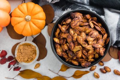 pumpkin seeds in a black bowl next to chocolate powder and an orange pumpkin