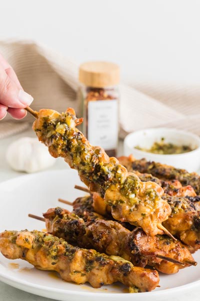 A hand holding a cooked chicken kebab over a plate with garlic, red pepper flakes and sauce in the background.