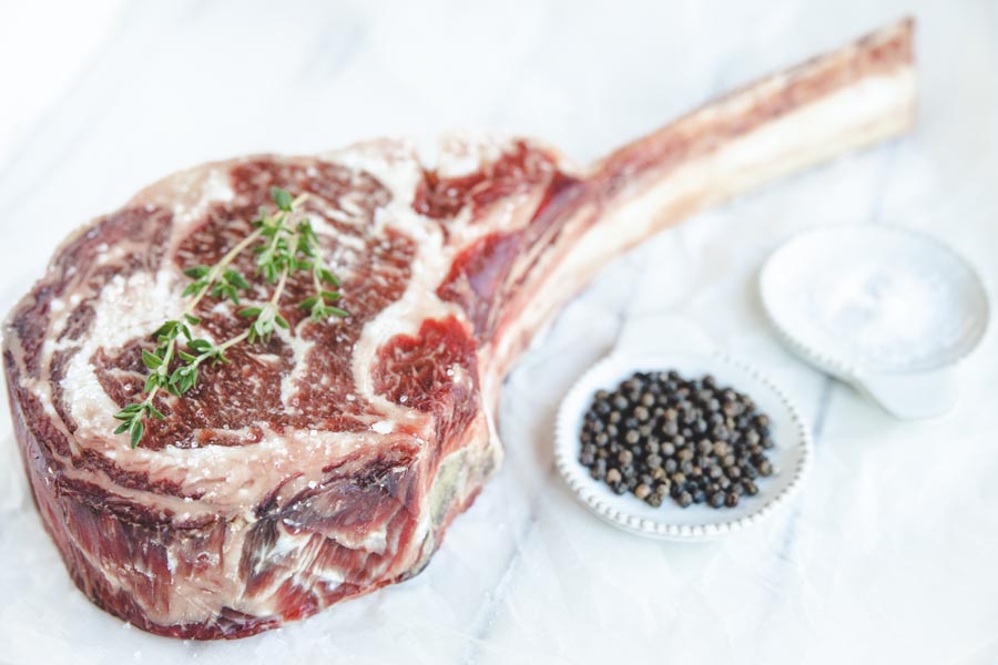 ribeye steak with a long bone next to a dish of salt and peppercorns