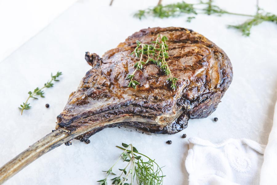 thick juicy tomahawk steak one in on a board with peppercorns and herbs scattered