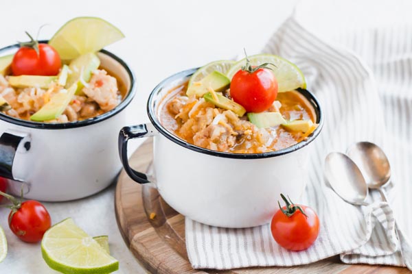 two bowls of low carb chicken & rice mixture in white enamel mugs on a striped linen towel with cherry tomatoes scattered around