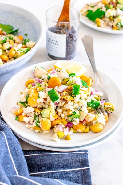 A small white plate holding a portion of a lower carb version of the Jennifer Aniston salad made famous from TikTok. The plate is filled with cauliflower rice, lupini beans, cucumber, feta and red onion. A jar of black peppercorns sits behind.