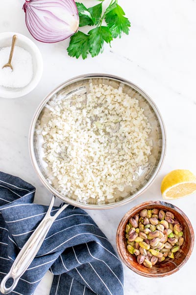 A skillet with cooked cauliflower rice is next to a bowl of pistachios, half a lemon and half a purple onion.