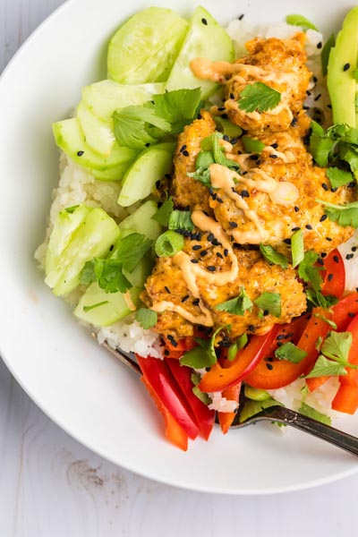 Crispy pieces of chicken topped with bang bang sauce over cauliflower rice and next to cucumber.