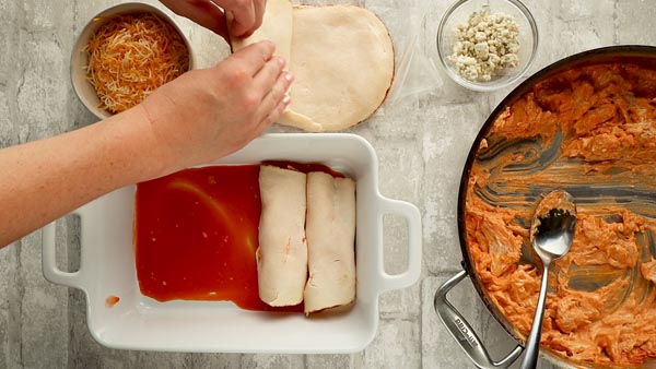 a couple rows of enchiladas in a casserole dish while grabbing sliced deli meat