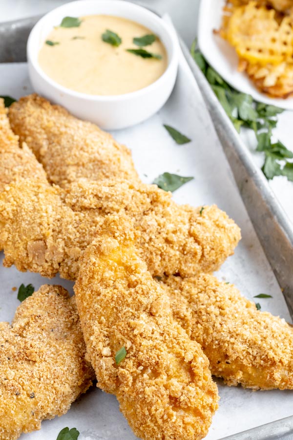 a bunch of crunchy breaded chicken tenders on a baking tray and sprinkled with parsley with a bowl of sauce behind them