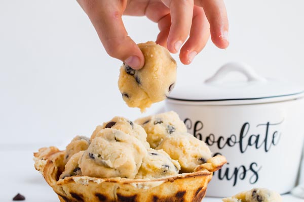 a child's hand holding a cookie dough bite over a bowl of cookie dough balls