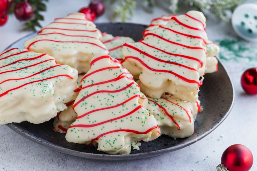 A pile a small cakes shaped like Little Debbie's Christmas Trees on a gray plate with Christmas bulbs nearby.