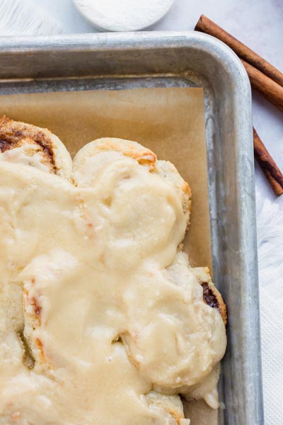 Baked and frosting cinnamon rolls on a tray with cinnamon sticks nearby.