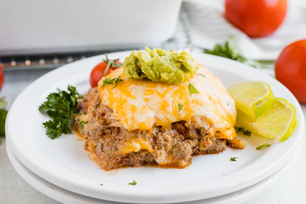 a slice of keto enchilada casserol on a white plate with tomato and lime slices in the background