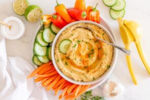 A platter with hummus, fresh veggies, salt, limes and garlic nearby.