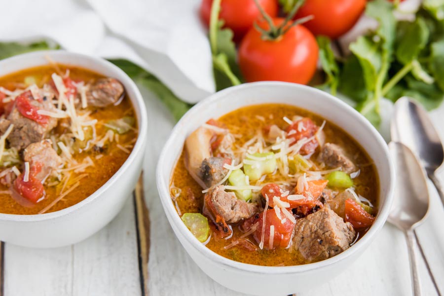 two bowls of beef stew and veggies for dinner next to tomatoes and kale