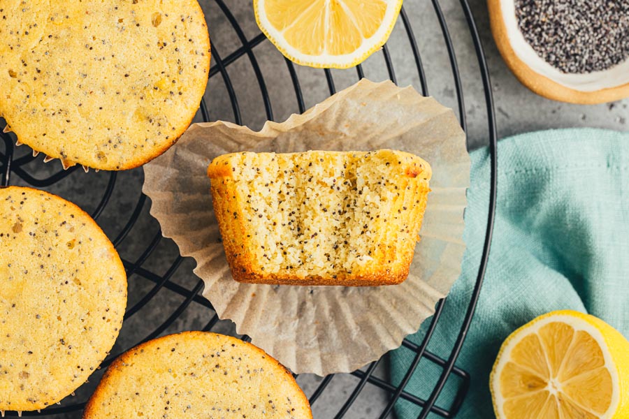 A bite out of lemon poppyseed muffins on a liner next to more muffins on a wire rack.