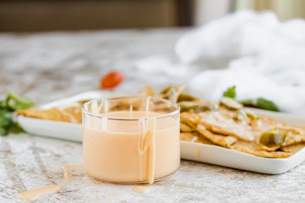 nacho cheese dripping down the side of a bowl of cheese with a plate of nachos in the background