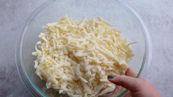 A hand holding a bowl of shredded mozzarella cheese.