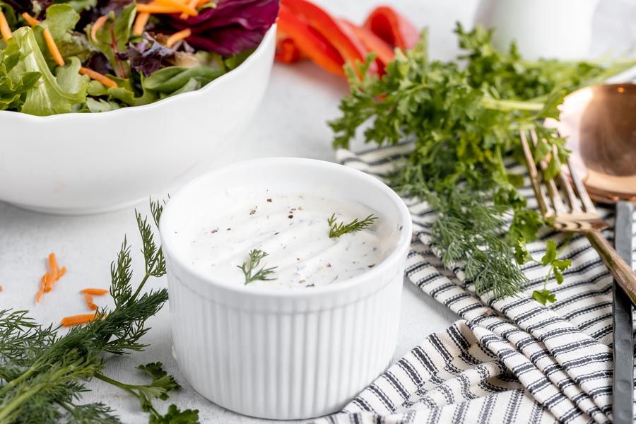 dressing in a small bowl with a gold spoon and fork to the right along with fresh dill and lettuce and a bowl of salad sites behind