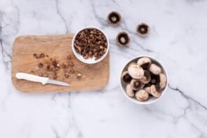 A cutting board with diced mushroom stems and a knife next to a bowl of mushroom caps.