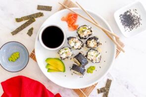 Sushi on a white plate with pickled ginger, avocado, a bowl of soy sauce, wasabi and chopsticks on the plate as well.