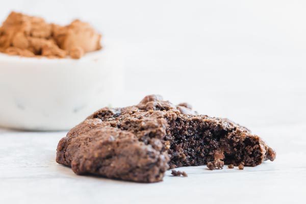 a half bitten chocolate cookie with a bowl of cocoa powder in the background