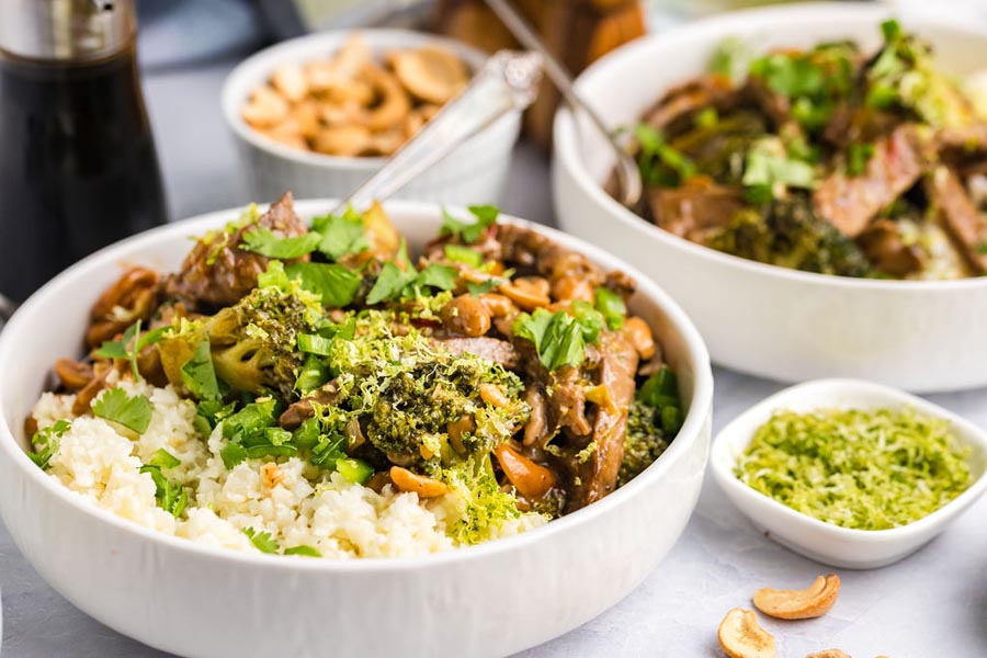 Two bowls of broccoli and cashew beef over cauliflower rice with lime zest nearby.