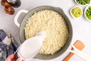 Pouring coconut milk into a skillet with riced cauliflower.