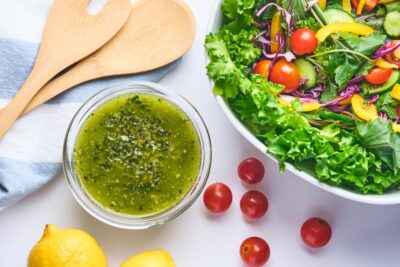 Green pesto dressing sits next to a bowl of salad, fresh tomatoes, lemons and salad tongs.