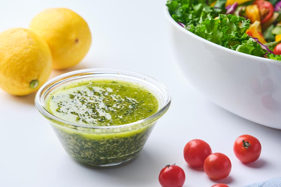 A small bowl of salad dressing sitting to the side of a bowl of salad, tomatoes and lemons.