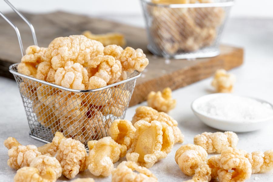 A mini fry basket filled with pork rinds and some scattered around next to a pinch bowl of salt.