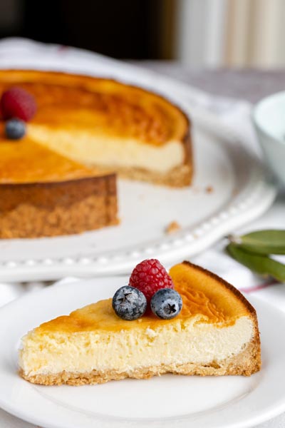 A small slice of cheesecake on a plate topped with blueberries and a raspberry. A full cheesecake sits behind.