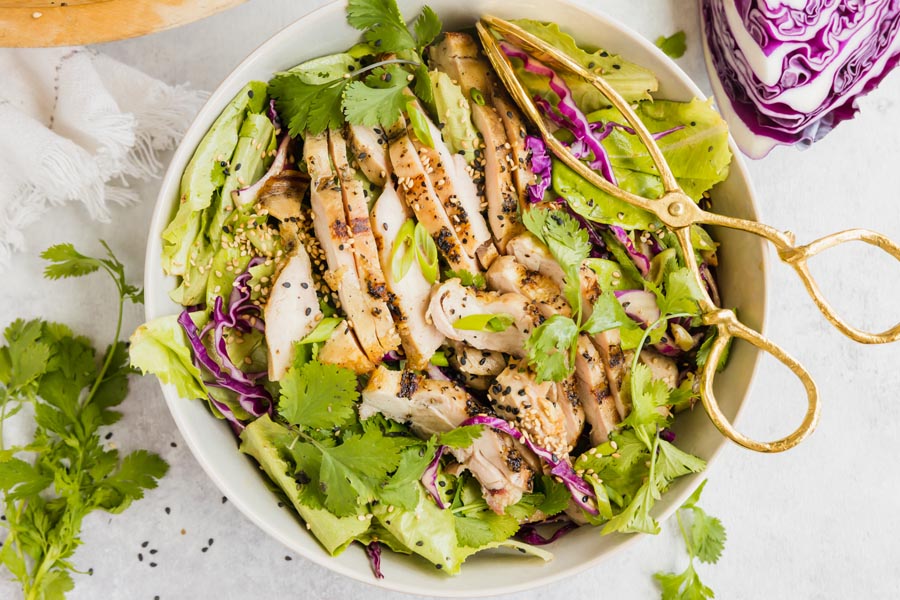 top down view of a chicken sald with cilantro green onions and purple cabbage with a pair of tongs on top