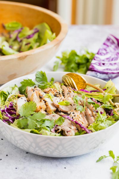 a bowl of salad with tongs sits in front of a bigger salad bowl and a red cabbage wedge