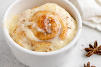 A warm cinnamon roll in a mug topped with cream cheese icing that is melting down into the cinnamon roll. Star anise is beside the mug.