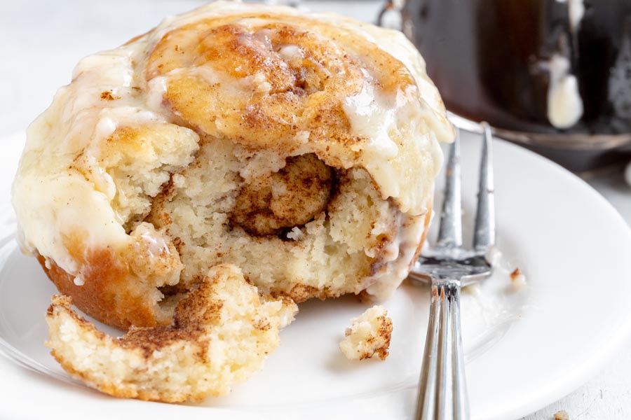 A bite missing from a cinnamon roll on a white plate with a fork next to the cinnamon roll. You can see the layers of buttery cinnamon inside the roll.